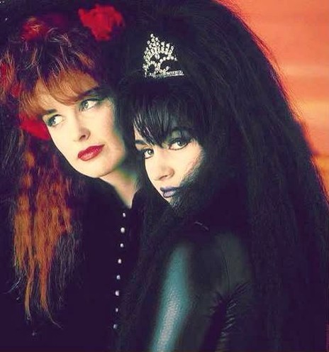 Close up photo ina  studio. Jill has red lipstick, crimped red hair and a hairband with red plastic roses. Rose stands next to her in a black PVC catsuit with black crimped hair and a diamante tiara, with purpley blue lipstick.