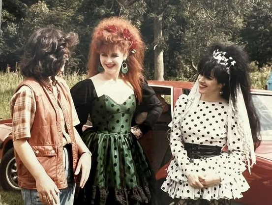 Outdoor photo, unposed, with Jill and Rose smiling at an unknown person. It's a sunny day, on the left is a person turned away from camera with long dark hair, wearing a red body warmer, checked shirt and blue jeans. In the centre is Jill Bryson with red back combed hair and a green strapless dress with black polka dots and black velevet sleeves. On the right is Rose McDowall in a white dress with black polka dots and ra-ra skirt.