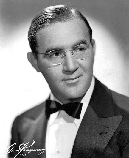 Black and white photo of Benny Goodman, a slim man with slicked back light hair in a tuxedo jacket, bowtie, and wire rimmed glasses. He's looking off camera and looks slightly bemused.