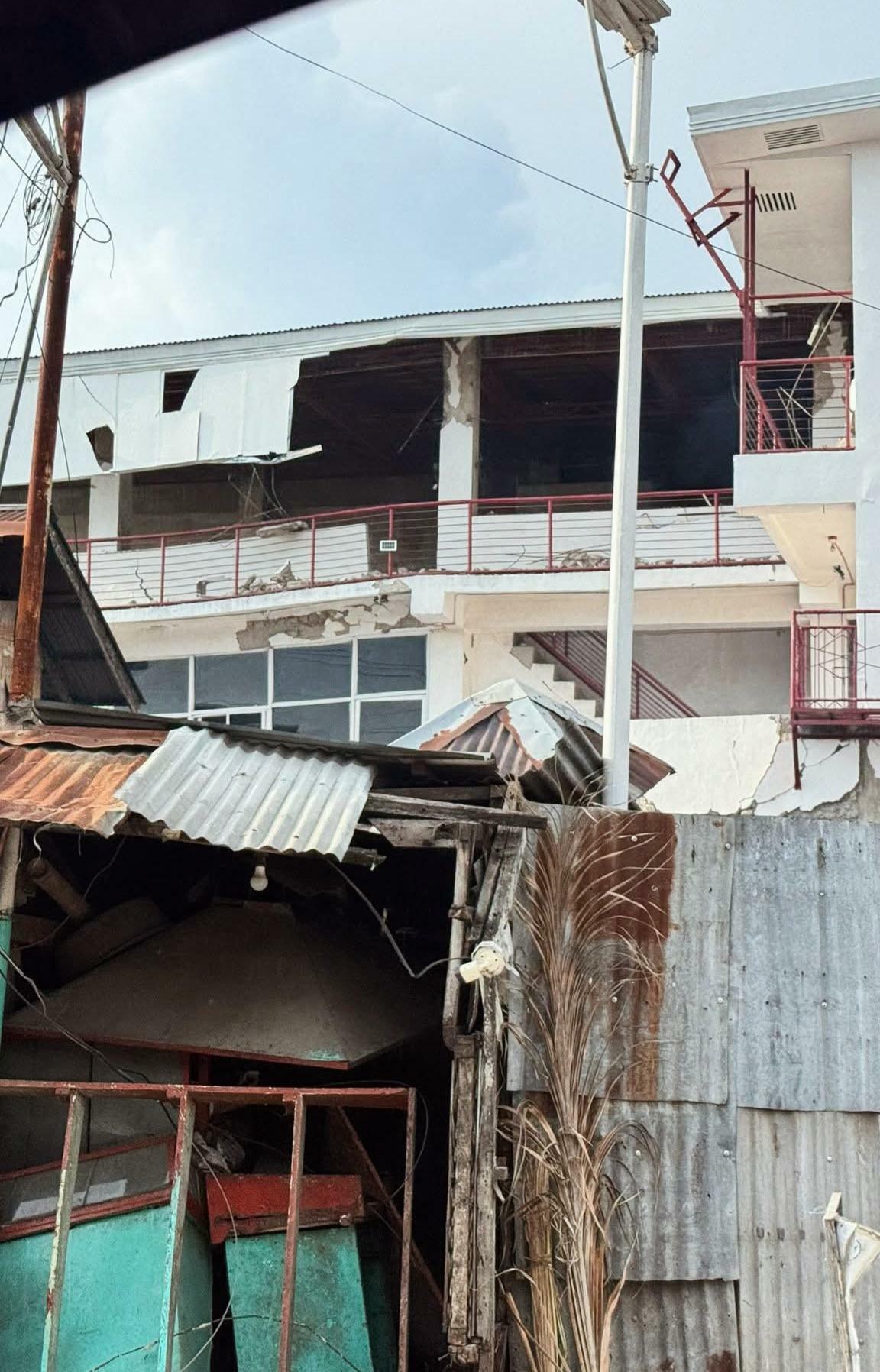 Close-up view of a severely damaged, multi-story white building, likely a commercial or institutional structure, in the aftermath of the Cebu earthquake. The top floor's exterior wall has completely collapsed, revealing the wooden roof structure and interior concrete columns. Cracks are visible on the remaining white and red balconies and walls.